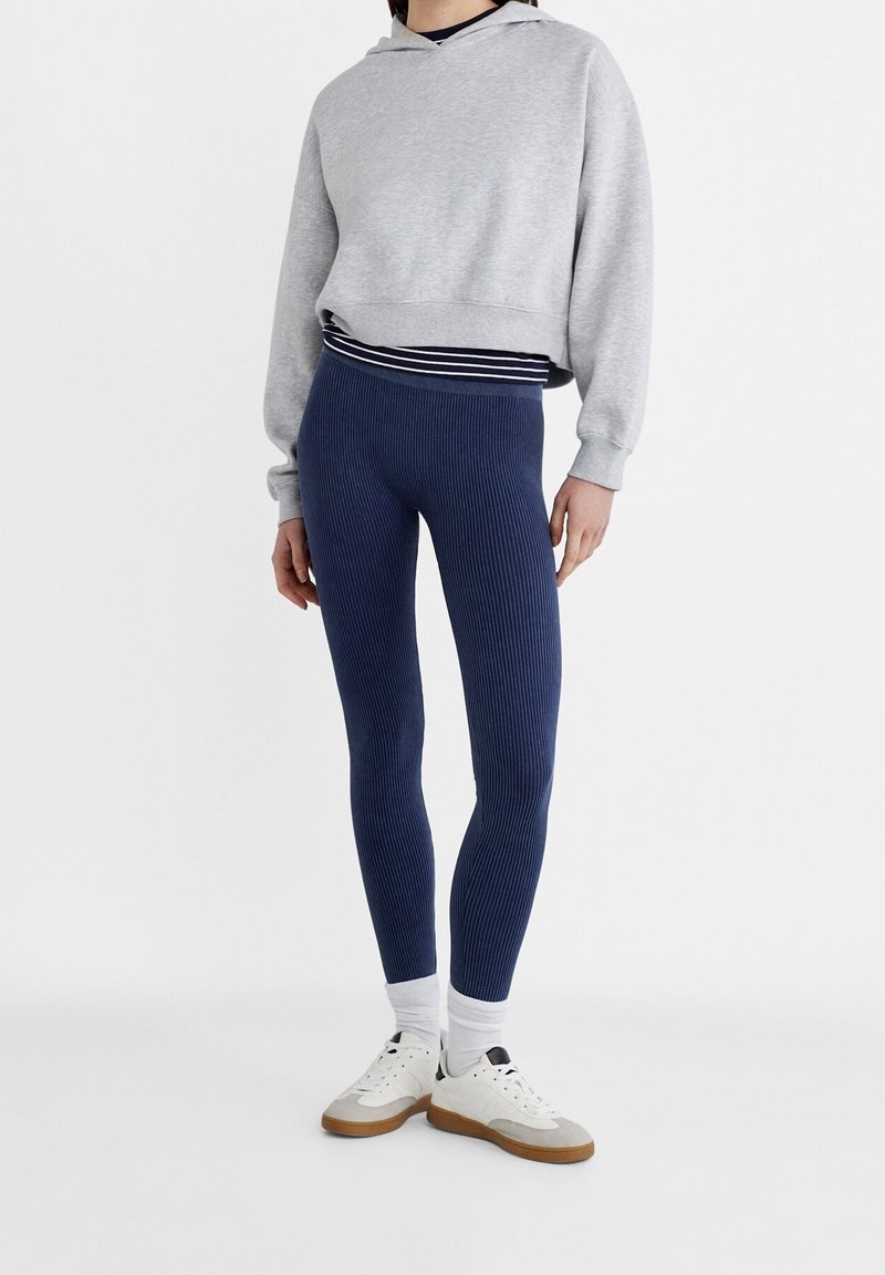 Sudadera recortada gris claro con capucha, leggings acanalados azul marino y zapatillas blancas con suelas marrones. Capa con una camiseta de rayas debajo.