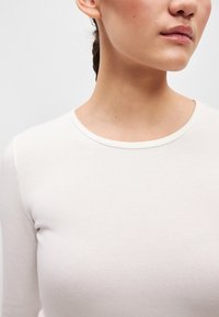 Hosszú ujjú, kerek nyakú felső puha, krémszínű pamutból. Sima textúrájú, minimális varrásokkal, amely egy egyszerű, testhezálló fazont mutat be.