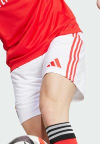 Rotes Sporttrikot mit Mesh-Struktur, kombiniert mit weißen Shorts mit roten Streifen und einer schwarz-roten gestreiften Socke, Fußball am Fuß.