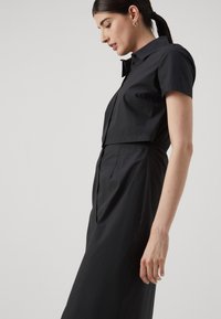 Abito nero a camicia realizzato in tessuto leggero, con maniche corte, un top aderente e un gonnellino morbido, dotato di bottoni frontali e vita elastica.