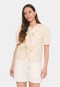 Quiltana prsluk u svijetloj krem boji s laganim zelenim uzorkom srca, s vezanjem sprijeda i napuhanim kratkim rukavima, nosi se preko svijetlih šortseva.