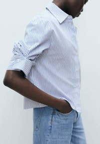 Chemise à rayures bleues et blanches avec boutons, col, manches retroussées et coupe décontractée. Associée à un jean denim bleu clair.