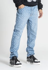 Vaqueros de mezclilla azul claro con un patrón de logo repetido, corte recto y textura ligeramente deslavada. Combinados con zapatillas de plataforma en blanco y negro.
