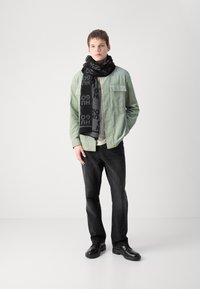 Jachetă din corduroy verde, cămașă albă și blugi negri. Șal negru cu modele de logo. Pantofi negri cu talpă groasă. Fundal neutru.
