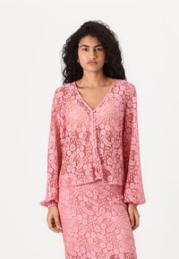 Rosa spetsblus med V-ringning, långa ärmar och knäppningar. Har blommönster och genomskinlig textur. Kompletterar en matchande kjol.