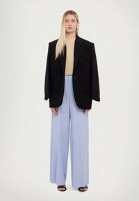 Sieviete ar gariem blondiem matiem, tērpusies melnā pārāk lielā žaketē, gaišā krēmkrāsas augšpusē, platās gaiši zilās biksēs un melnās atvērtajās papēžkurpēs, stāvot uz baltas fona.