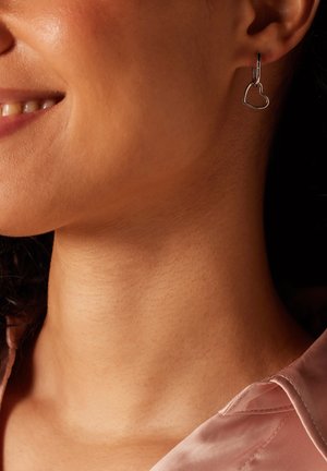 Boucle d'oreille en argent en forme de cœur sur le lobe, avec un design épuré et une finition lisse et polie. Le teint de la peau est chaud.