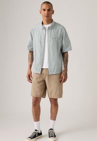 Korte mouwen groen-wit gestreept shirt met een borstzak, gecombineerd met lichtbeige shorts en zwarte sneakers met witte zolen.