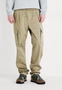 Beige cargo bukser med flere sidelommer, elastisk talje og en løs pasform; parret med grønne og brune sneakers.