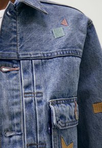 Giubbotto di jeans realizzato in tessuto blu, con cuciture a contrasto, toppe ricamate geometriche in blu, rosa e beige, e una tasca a pattina.