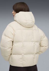 Giacca puffer crema con cappuccio, caratterizzata da un design trapuntato e una vestibilità oversize. Texture liscia, lunghezza corta e polsini elasticizzati.