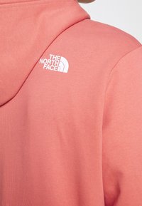 Sudadera melocotón hecha de tela suave, con un logo blanco de The North Face en el hombro izquierdo superior. Textura suave con capucha.