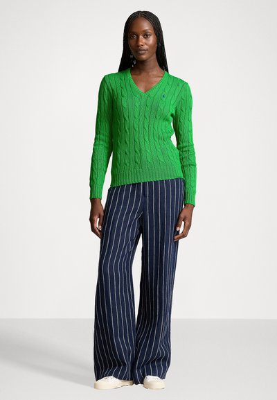 Pull en maille torsadée vert avec un décolleté en V, associé à un pantalon large rayé bleu marine. Le mannequin a les cheveux tressés et porte des chaussures blanches.