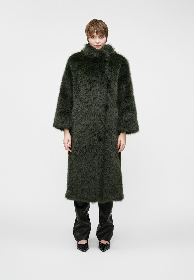 Długi, ciemnozielony futrzany płaszcz z wysokim kołnierzem, szerokimi rękawami i gładką teksturą. Noszony na czarnych skórzanych spodniach oraz spiczastych czarnych butach.
