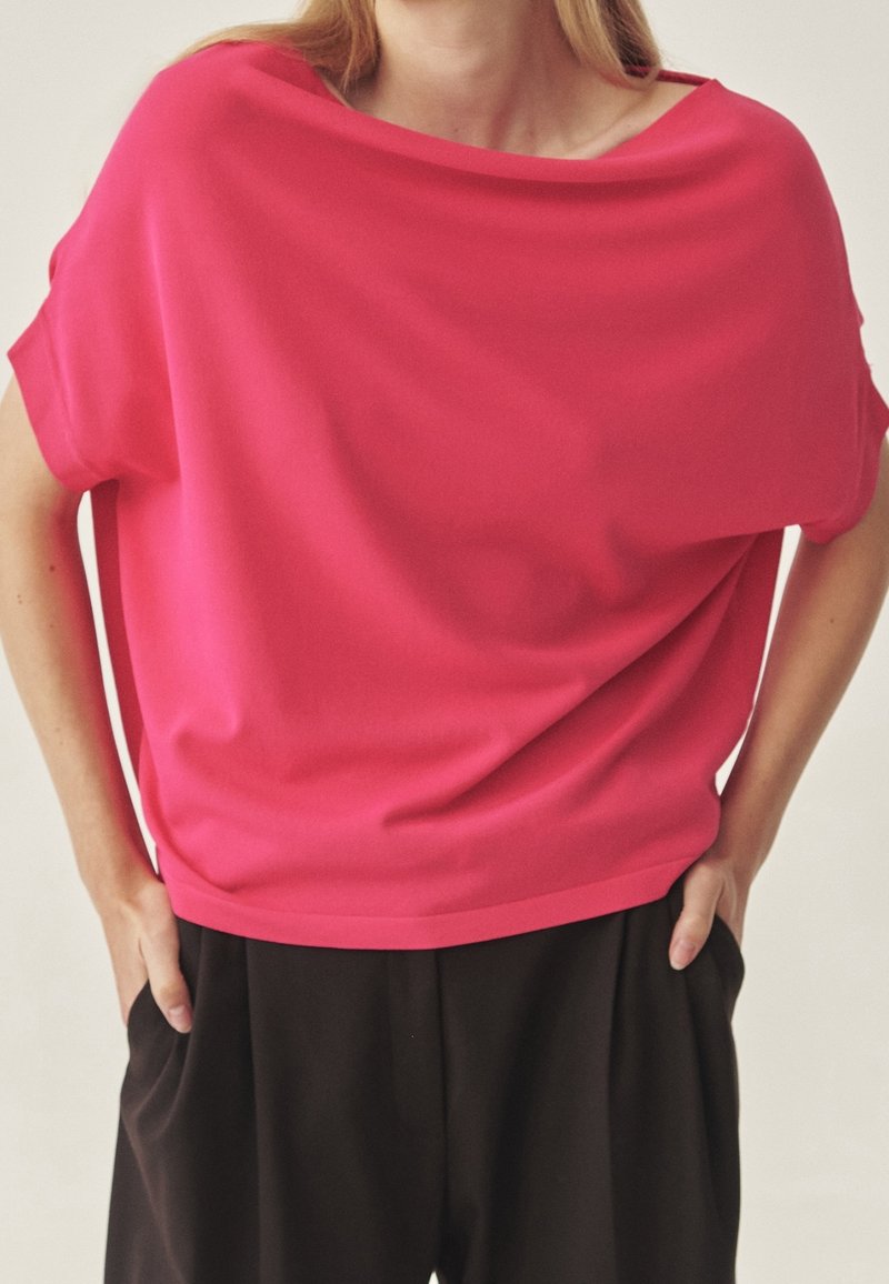 Haut oversize fuchsia en tissu lisse, avec un décolleté large et des manches courtes, associé à un pantalon noir ample.
