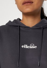 Personne portant un sweat à capuche gris foncé avec des cordons de serrage, arborant un logo blanc "ellesse" centré sur la poitrine.