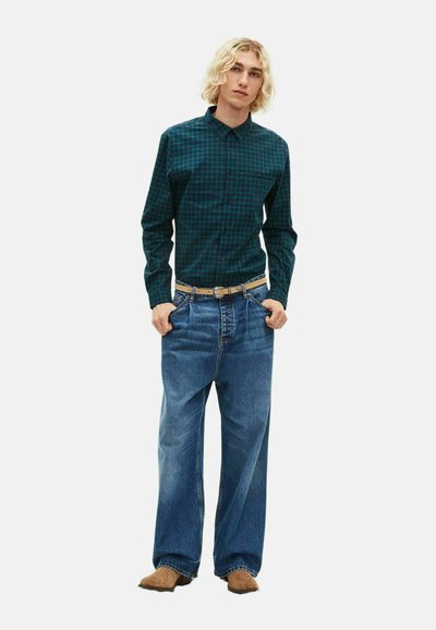 Camicia a quadri verde e nera, jeans in denim azzurro chiaro a vestibilità morbida, cintura beige e scarpe marroni, in piedi davanti a uno sfondo bianco.