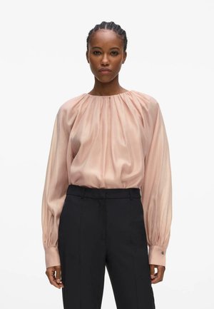 Žena s vrkočmi nosí bledoružovú blúzku s dlhými rukávmi a naberanými ramenami, zastrčenú do čiernych nohavíc s vysokým pásom, stojacu na bielom pozadí.