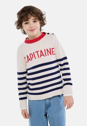 Enfant aux cheveux bouclés portant un pull blanc avec le texte rouge "CAPITAINE", des rayures bleu marine, un col rouge et un jean bleu, sourire léger.