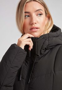 Chaqueta acolchada negra con cuello alto, frontal con cremallera y capucha ajustable. Presenta secciones acolchadas y tela texturizada para proporcionar calidez.