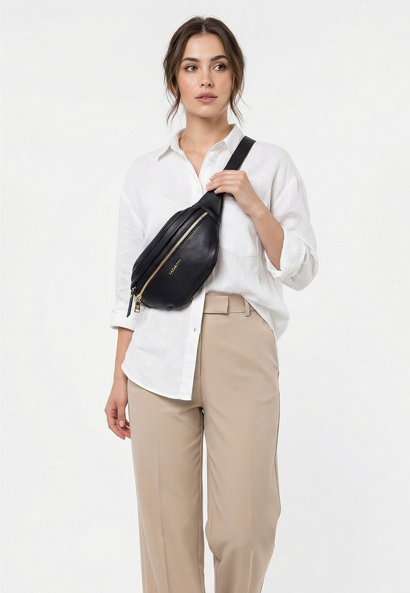 Femme portant une chemise blanche à boutons et un pantalon beige, portant un sac bandoulière en cuir noir avec une fermeture éclair dorée sur la poitrine.