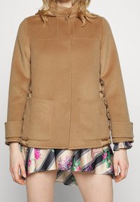 Brauner Wollmischungsmantel mit hohem Kragen, Seitentaschen und gefältelten Akzenten an den Ärmeln. Getragen über einem gemusterten, ausgestellten Blumrock.