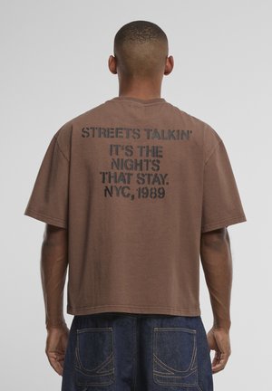 Hombre con camiseta marrón con texto negro "STREETS TALKIN' IT'S THE NIGHTS THAT STAY. NYC, 1989" en la espalda, combinado con vaqueros azules.