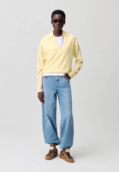 Persona con gafas de sol oscuras, camisa polo de manga larga color amarillo pálido, jeans holgados de color azul claro y zapatillas marrones, de pie contra un fondo liso.