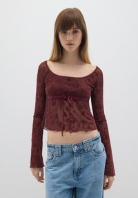 Top lungo trasparente bordeaux con maniche lunghe, motivo floreale, scollo a barca e dettaglio con cordoncino in vita, abbinato a jeans azzurri chiari.