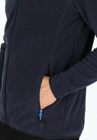 Marineblaue Fleecejacke mit durchgehendem Reißverschluss, seitlichen Taschen mit blauen Akzenten und langen Ärmeln mit taillierten Bündchen. Weiche Textur und lässiges Design.