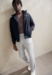 Giacca leggera della marina con cappuccio e logo sul petto, abbinata a pantaloni della tuta grigi con logo, indossata con scarpe da ginnastica bianche.