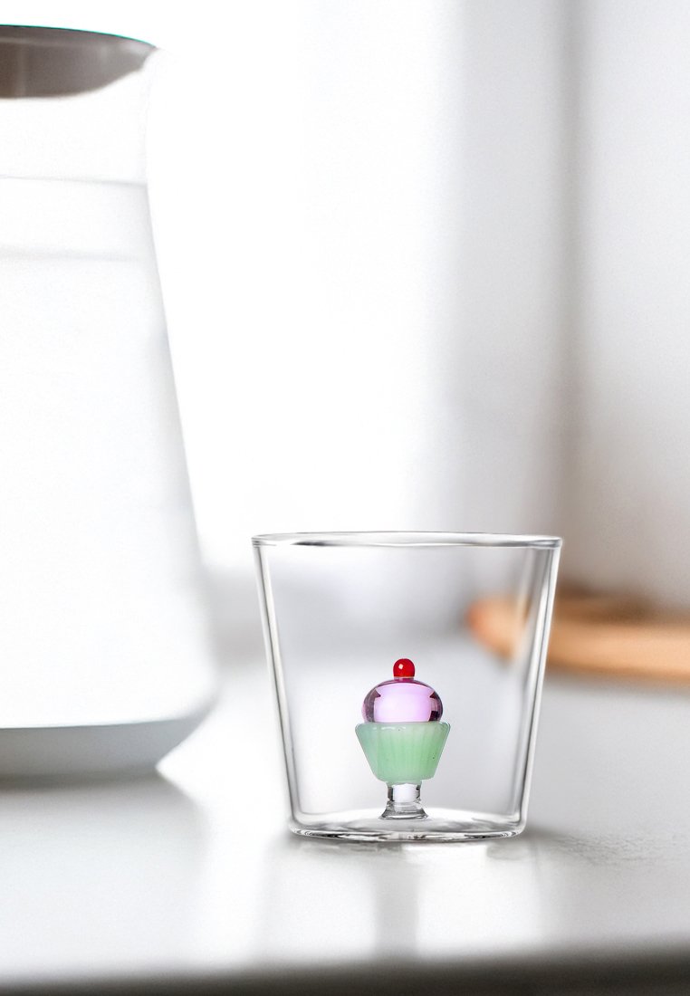 Przezroczysta szklanka zawierająca dekoracyjną figurkę babeczki z czerwoną wiśnią na wierzchu, fioletowymi i zielonymi warstwami, gładką teksturą oraz zaokrąglonym kształtem.