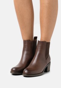 Bottines en cuir marron avec un panneau latéral élastique texturé, bout rond et un talon cubique. La semelle est légèrement surélevée.