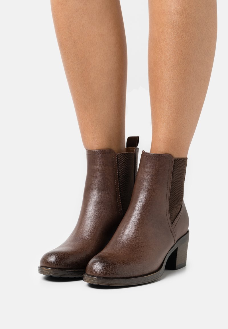 Bottines en cuir marron avec un panneau latéral élastique texturé, bout rond et un talon cubique. La semelle est légèrement surélevée.
