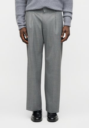 Άνδρας που φοράει γκρι ραμμένα παντελόνια, μαύρα επίσημα παπούτσια και ανοιχτό γκρι χνουδωτό πουλόβερ, στέκεται μπροστά σε μια απλή φόντο.