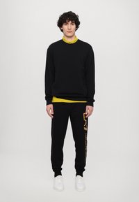 Felpa nera con tasca, indossata sopra una maglietta gialla. Pantaloni sportivi neri con stampa del logo laterale e sneakers bianche. Tessuto liscio in tutto.