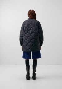 Person with long braids wearing a black quilted jacket, blue knee-length shorts, black leggings, and black mid-calf boots, shown from behind.