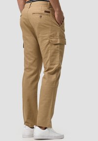 Pantalons cargo beiges en coton, avec poches latérales, une poche arrière avec bouton et une coupe droite. Associés à des baskets blanches.