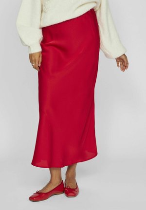 Jupe maxi en satin rouge avec taille élastique, texture lisse, associée à des chaussures plates rouges avec des accents de nœud.