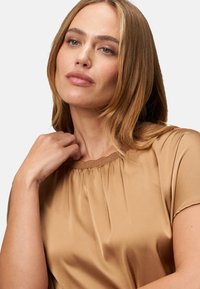 Bežový top, hladká saténová textura, krátké rukávy, nabíraný výstřih, jemný lesk. Modelka má světle hnědé vlasy, minimální make-up, neutrální pozadí.