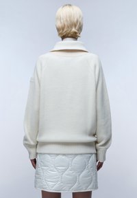 Witte gebreide trui met een hoge kraag en geribde mouwen, gecombineerd met een glanzende witte gewatteerde rok met een golvend patroon.