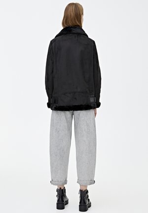 Persoon met lichtbruin haar die een zwarte schapenvachtjas draagt, losse grijze opgerolde jeans en zwarte enkellaarsjes, staand met de rug naar de camera.