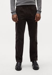 Pantaloni in velluto a coste di colore marrone scuro con righe verticali, dotati di chiusura a bottone, tasche laterali e taglio dritto, abbinati a scarpe nere.