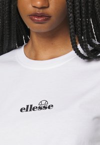 Bílá bavlněná tričko s černým logem "ellesse" na hrudi, kulatým výstřihem a hladkým, nezdobeným povrchem.