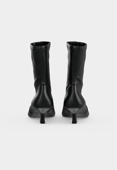 Paire de bottes en cuir noir mi-mollet avec talons étroits, vues de dos sur un fond clair uni.