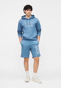 Blå hoodie i bomullsblandning med framficka och vitt logo, tillsammans med matchande shorts med sidotryckt logo och vita sneakers.