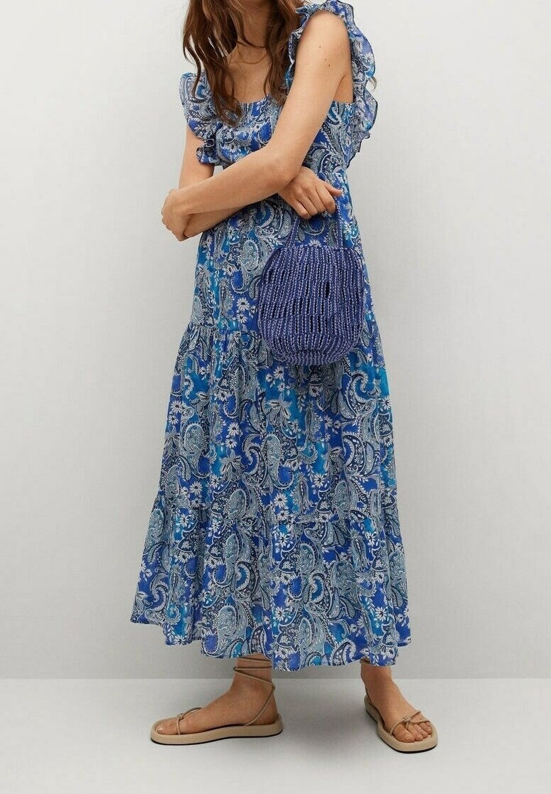Femme portant une robe bleue à manches volantées avec motif cachemire, des sandales beiges à brides et tenant un sac à main rond en osier bleu marine.