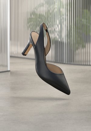Scarpa con tacco alto slingback in pelle nera con punta a punta, sospesa sopra un pavimento liscio in cemento e uno sfondo moderno con barre metalliche verticali.
