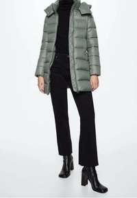 Frau trägt eine hellgraue Steppjacke über einem schwarzen Rollkragenpullover, schwarze Hose und schwarze Stiefeletten mit Absatz, steht vor einem weißen Hintergrund.