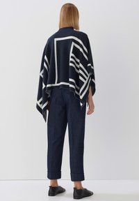 Navy-blaues Poncho mit weißen geometrischen Streifen, aus weichem Stoff. Kombiniert mit dunklen Jeans und schwarzen Schuhen.